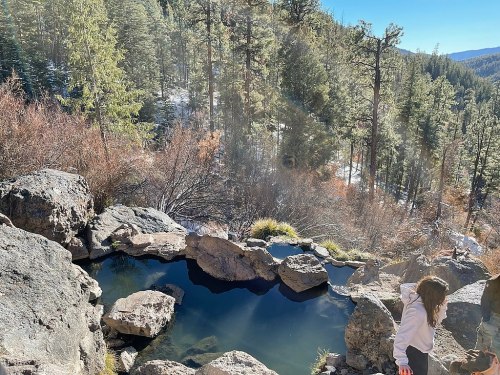 A serene natural hot spring surrounded by rocks and trees, with a person standing nearby in a sunny landscape.