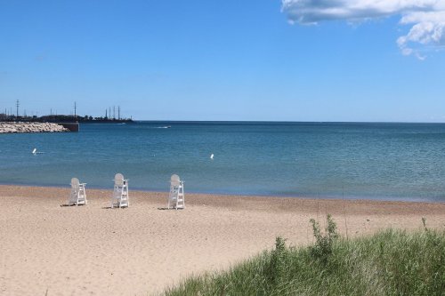 Three white beach chairs on a sandy shore, overlooking a calm blue lake under a clear sky.