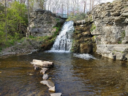 A serene waterfall cascades over rocky cliffs into a calm pool, surrounded by lush greenery and a wooden bridge above.