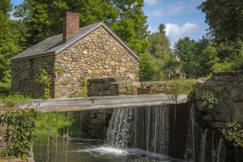 A stone building beside a small waterfall, surrounded by lush greenery and a clear blue sky.