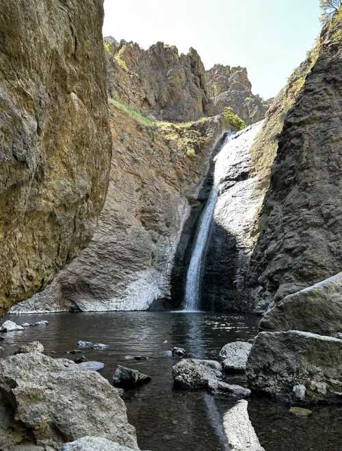 A waterfall cascades down rocky cliffs into a serene pool, surrounded by rugged terrain and clear blue skies.