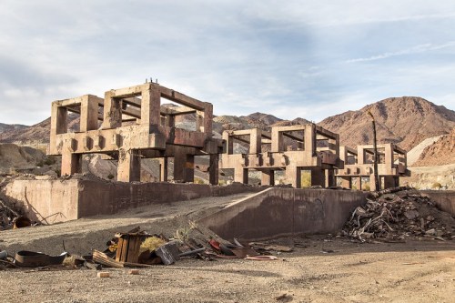 Abandoned concrete structures in a desert landscape, surrounded by debris and mountains in the background.