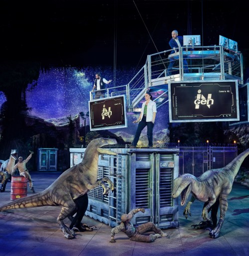 A dramatic stage scene with actors and dinosaurs, featuring technology displays and a vibrant cosmic backdrop.