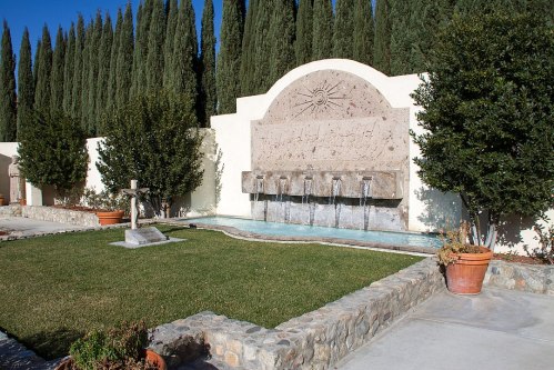 A stone fountain with flowing water, surrounded by greenery and cypress trees, in a serene garden setting.
