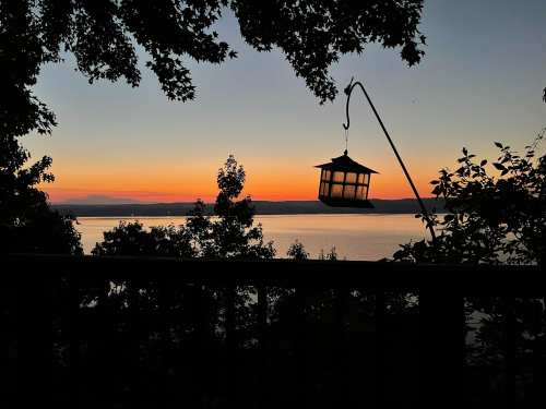 A serene sunset over a lake, with silhouettes of trees and a lantern hanging from a post in the foreground.