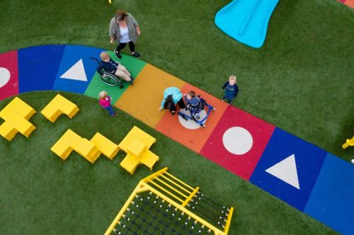 A colorful playground with children playing, including one in a wheelchair, and a caregiver nearby.