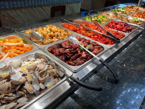 A buffet display featuring various seafood and meat dishes in metal trays on a dark countertop.