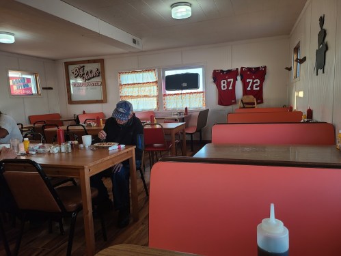 A cozy diner interior with wooden tables, a person eating, and sports jerseys hanging on the wall.