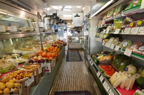 A vibrant market aisle filled with fresh fruits, vegetables, and various goods, with wooden floors and bright lighting.
