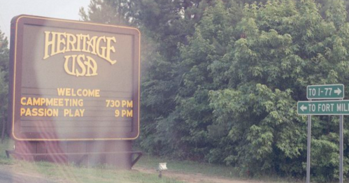 Sign for Heritage USA welcoming visitors to a camp meeting at 7:30 PM and Passion Play at 9 PM, with directional signs.