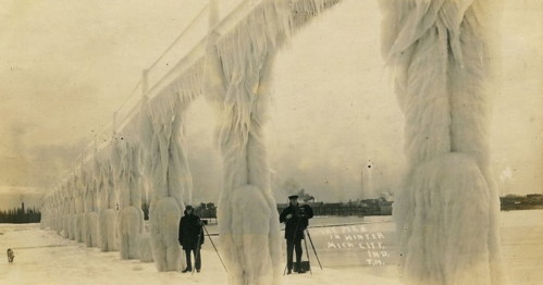 Two people stand on a frozen bridge covered in ice, with a snowy landscape in the background.