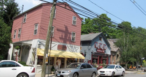 A quaint street scene featuring a pink building, a blue shop, and parked cars under power lines on a sunny day.