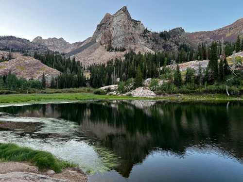 A serene lake reflects towering mountains and lush trees under a clear sky, creating a tranquil natural scene.