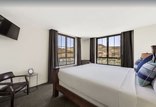 Bright hotel room with a large bed, two windows, a chair, and a TV, offering a view of the surrounding landscape.