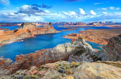 A stunning landscape of colorful rock formations and a blue lake under a bright sky with scattered clouds.