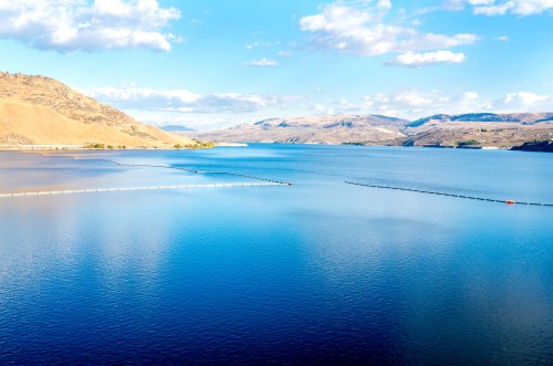 A serene lake surrounded by hills under a blue sky with scattered clouds, featuring calm waters and a floating barrier.