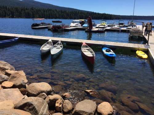 A serene lake scene with canoes and kayaks docked at a marina, surrounded by trees and mountains in the background.