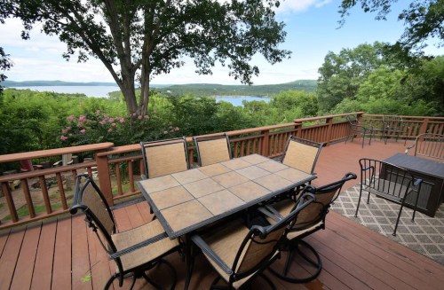 A wooden deck with a table and chairs overlooks a lush green landscape and a serene lake in the distance.