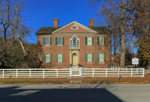 Historic brick house with green shutters, white fence, and clear blue sky in the background.