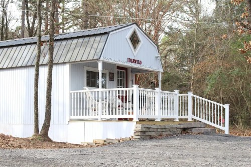 A small white cabin with a metal roof, featuring a porch and surrounded by trees.