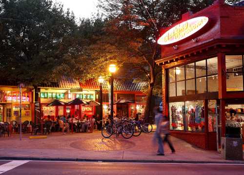 A vibrant street scene at dusk featuring shops, outdoor seating, and bicycles parked near a pizza restaurant.