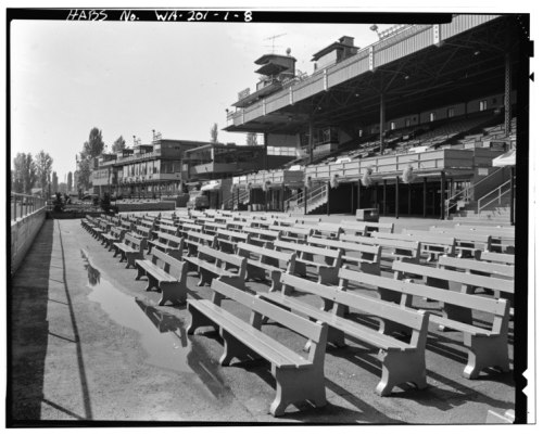 Empty grandstand with rows of benches, reflecting puddles on the ground, under a clear sky. Historical black and white photo.