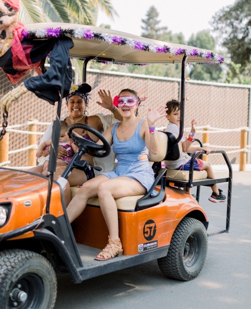 A group of three people, including a child, joyfully waving from an orange golf cart decorated with festive accessories.