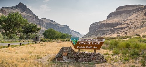 Sign for Owyhee River Wilderness Area, surrounded by rocky cliffs and grassy terrain under a hazy sky.