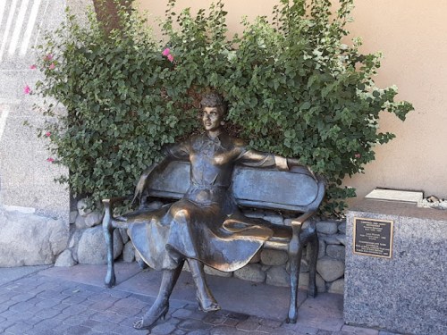 A bronze statue of a woman sitting on a bench, surrounded by greenery and flowers.