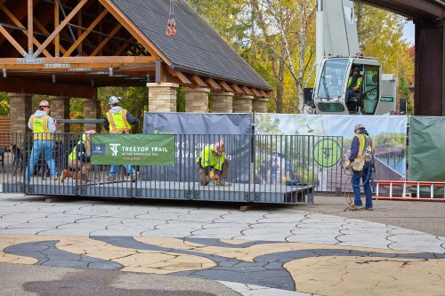 Construction workers in safety gear work on a platform near a building, with machinery and a banner for Treetop Trail visible.