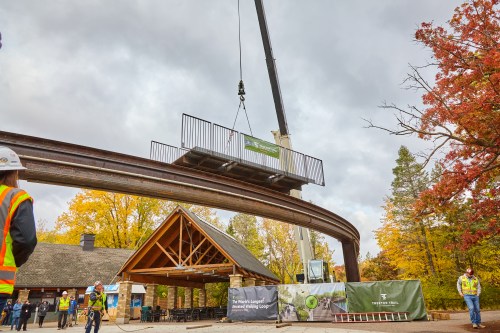 A crane lifts a structure above a building under cloudy skies, with workers and autumn trees in the background.
