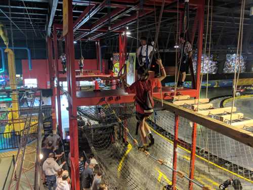 A busy indoor amusement park with people on a ropes course above a go-kart track. Safety harnesses are visible.