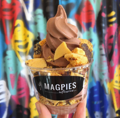 A cup of chocolate soft serve topped with crunchy pieces, held in front of a colorful graffiti background.