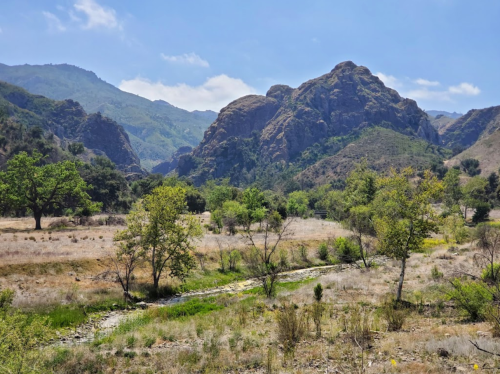 A scenic landscape featuring rolling hills, green trees, and a dry riverbed under a clear blue sky.