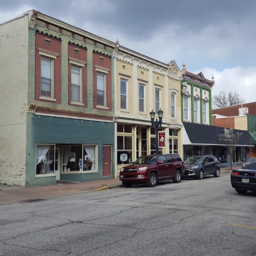 Historic buildings line a street, featuring colorful facades and parked cars under a cloudy sky.