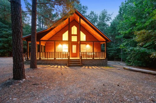 A cozy cabin with large windows, surrounded by trees, illuminated at dusk. Gravel path leads to the entrance.