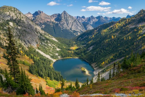 A scenic view of a mountain valley with a serene lake surrounded by lush forests and rugged peaks under a blue sky.
