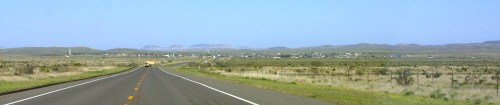 A long, straight road leads to a small town surrounded by mountains and open fields under a clear blue sky.