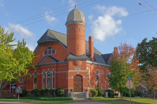 Historic red brick library building with a tower, surrounded by trees and a clear blue sky.