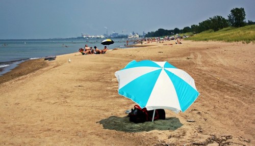 A sandy beach with colorful umbrellas, people relaxing, and a calm lake in the background under a clear sky.