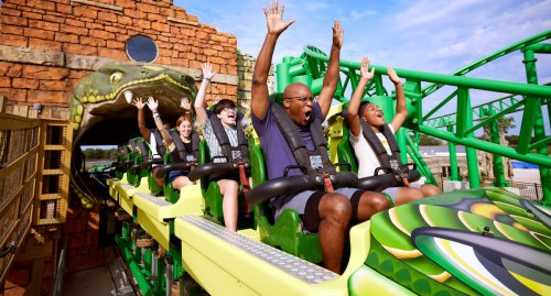 A group of excited riders with hands raised on a roller coaster, entering a themed tunnel with a snake design.