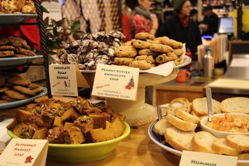 A display of baked goods including cookies, loaves, and hummus, with labels for each item at a cozy store.