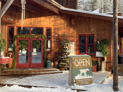A cozy wooden store with red doors, decorated for the holidays, and a sign reading "OPEN" in a snowy landscape.