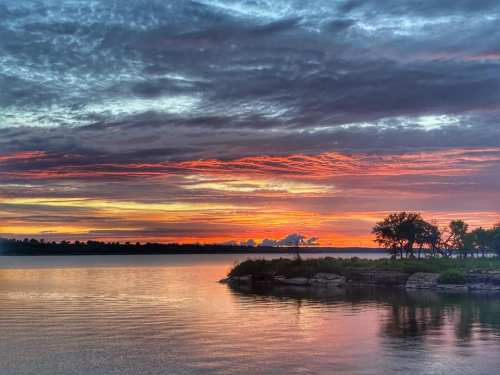 A serene sunset over a calm lake, with vibrant orange and purple hues reflecting on the water and silhouettes of trees.