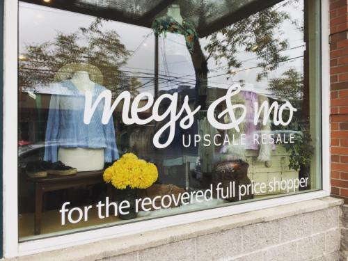 Storefront window displaying "megs & mo UPSCALE RESALE" with clothing and flowers, promoting upscale resale shopping.