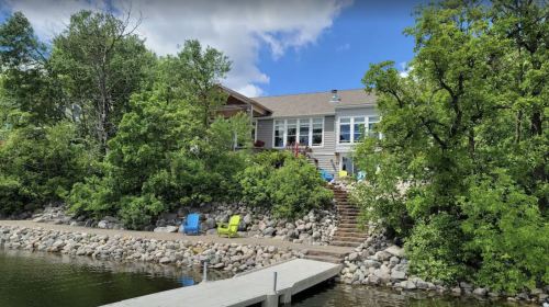 A lakeside house surrounded by trees, with a wooden dock and colorful chairs on the shore under a blue sky.