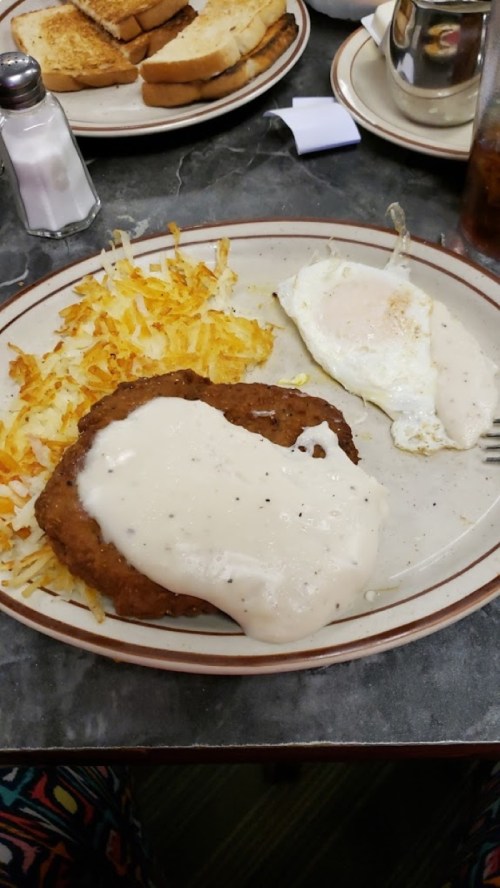 A plate with crispy hash browns, a fried egg, and a breaded meat patty topped with creamy gravy. Toast in the background.