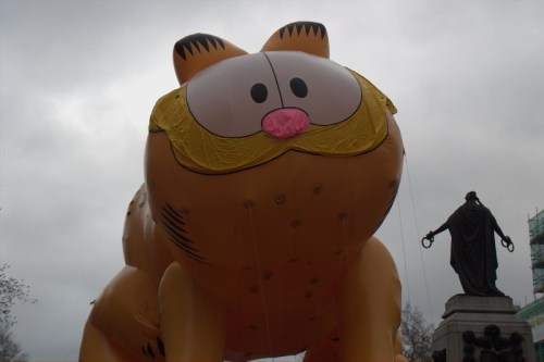 A large inflatable Garfield balloon against a cloudy sky, with a statue in the background.