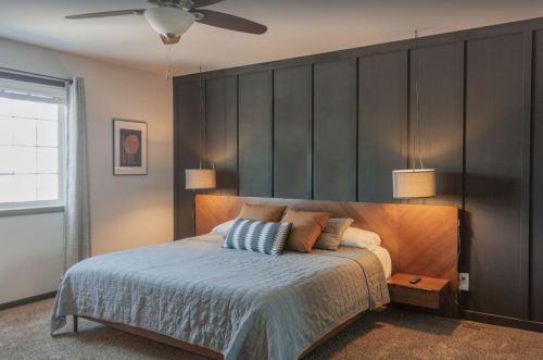 Cozy bedroom with a dark paneled wall, a bed with gray bedding, and modern pendant lights on either side.