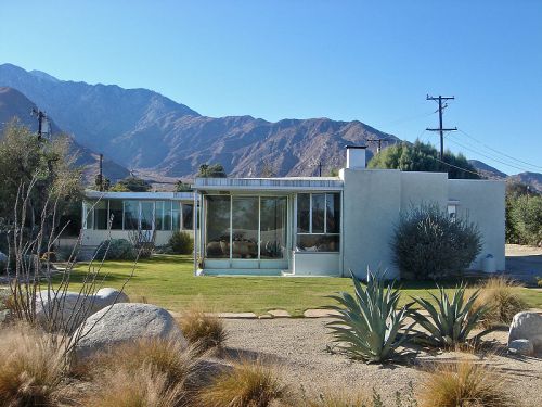 Modern house with large windows, surrounded by desert landscaping and mountains in the background under a clear blue sky.